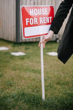 Close-up of a red 'House for Rent' sign on a grassy lawn, hand holding pole.